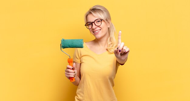 Mujer rubia sonriendo con orgullo y confianza haciendo la pose número uno triunfalmente, sintiéndose como una líder