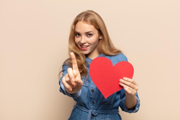 Mujer rubia sonriendo con orgullo y confianza haciendo la pose número uno triunfalmente, sintiéndose como una líder