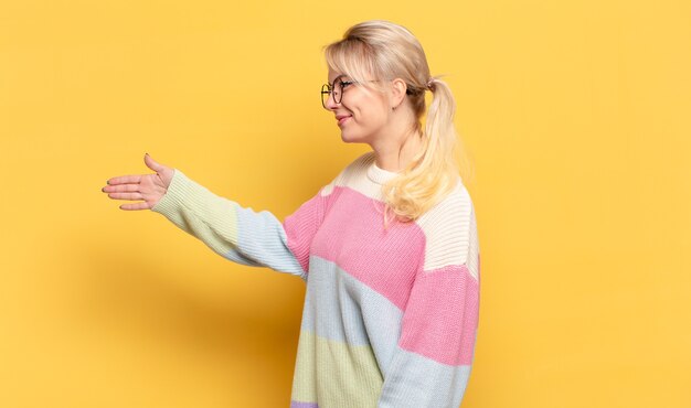 Mujer rubia sonriendo, saludándote y ofreciendo un apretón de manos para cerrar un trato exitoso, concepto de cooperación