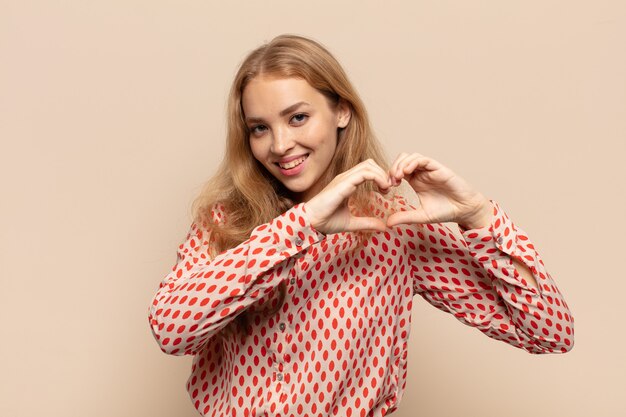 Mujer rubia sonriendo y sintiéndose feliz, linda, romántica y enamorada, haciendo forma de corazón con ambas manos