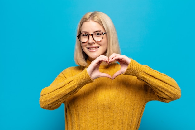 Mujer rubia sonriendo y sintiéndose feliz, linda, romántica y enamorada, haciendo forma de corazón con ambas manos