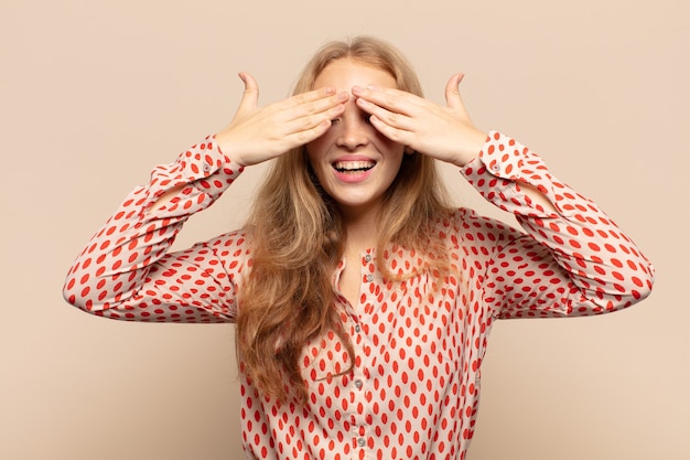 Mujer rubia sonriendo y sintiéndose feliz, tapándose los ojos con ambas manos y esperando una sorpresa increíble.