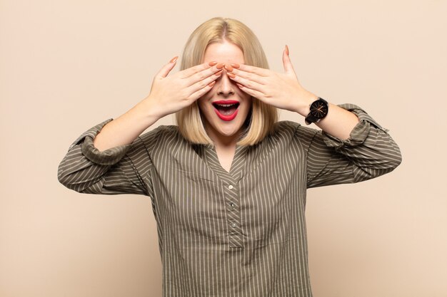 Mujer rubia sonriendo y sintiéndose feliz, tapándose los ojos con ambas manos y esperando una sorpresa increíble.