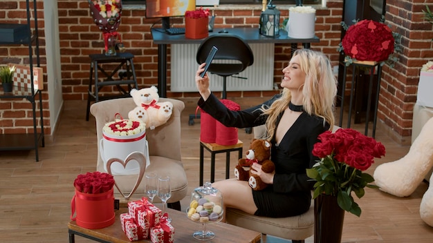 Mujer rubia sonriendo y tomando fotos con regalo de oso de peluche para la celebración del día de san valentín. Sorpresa romántica con ramo de flores y dulces regalos para celebrar la fiesta del amor.