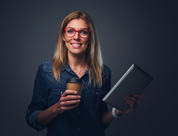 Una mujer rubia sonriente con anteojos sostiene una tableta y se lleva una taza de café.