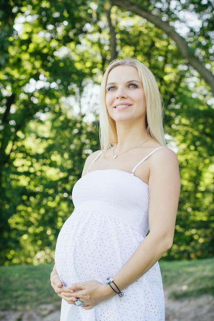 Mujer rubia sonriente embarazada en vestido blanco mirando hacia arriba y sosteniendo su gran barriga en el parque. Caminar al aire libre