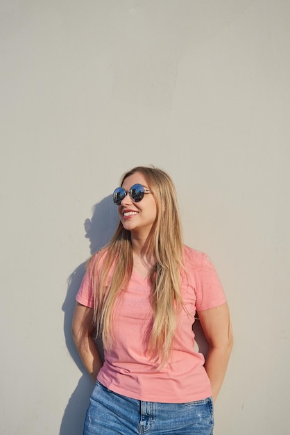 Mujer rubia sonriente feliz con gafas de sol en una camiseta rosa parada contra una pared gris en un día soleado