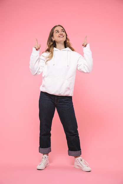 Mujer rubia sonriente de longitud completa con ropa casual apuntando y mirando hacia arriba sobre la pared rosa