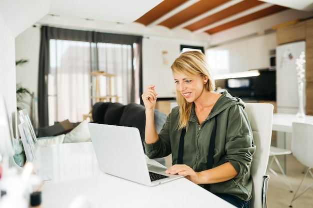 Mujer rubia sonriente de mediana edad usando una computadora portátil y tener una videollamada en una sala de estar