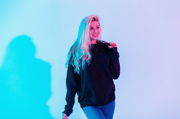 Mujer rubia sonriente de moda feliz en sudadera con capucha negra con jeans azul en estudio sobre fondo rosa neón multicolor