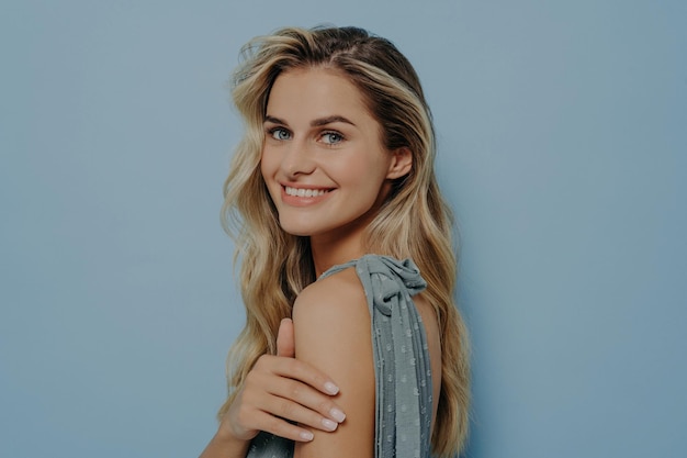 Mujer rubia sonriente con vestido azul mira por encima del hombro con una sonrisa elegante sosteniendo su propia mano aislada en el fondo azul