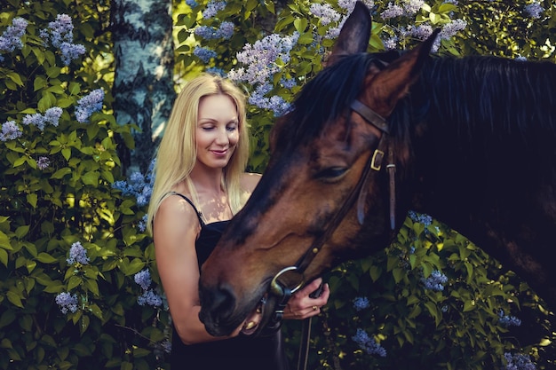 Mujer rubia sonriente con vestido negro y su caballo marrón.