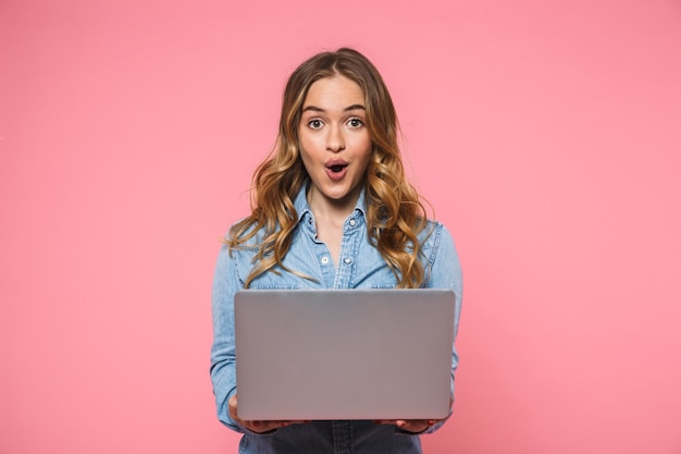 Mujer rubia sorprendida con camisa de mezclilla sosteniendo una computadora portátil y mirando al frente con la boca abierta sobre la pared rosa