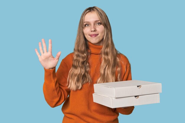 Foto mujer rubia sosteniendo cajas de pizza en el estudio sonriendo alegremente mostrando el número cinco con los dedos