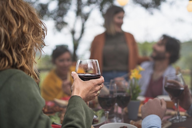 Mujer rubia sosteniendo una copa de vino tinto hablando con amigos en una mesa de picnic en el campo enfocándose en la mano de personas irreconocibles desenfocadas en el concepto de estilo de vida de degustación de vinos de fondo