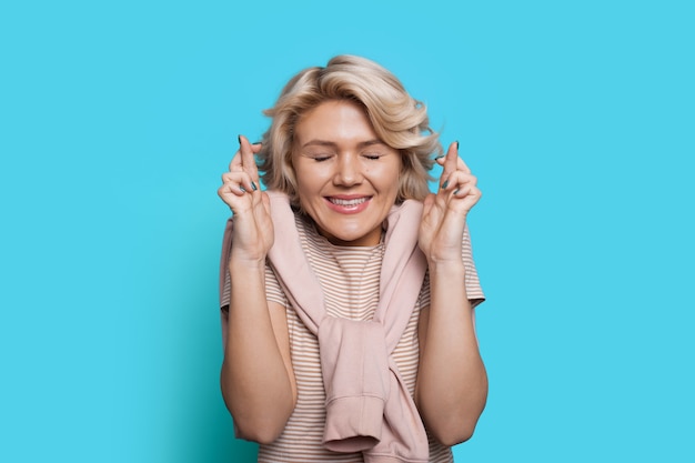 Mujer rubia sueña con algo cruzando los dedos y posando con los ojos cerrados en una pared azul