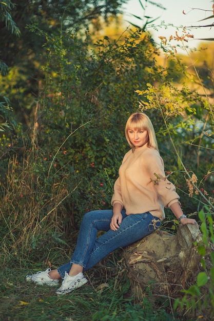 Una mujer rubia con un suéter de punto con un corte de pelo corto está sentada sobre un árbol caído en el fondo de un estanque en el bosque. Recreación al aire libre en el bosque de otoño