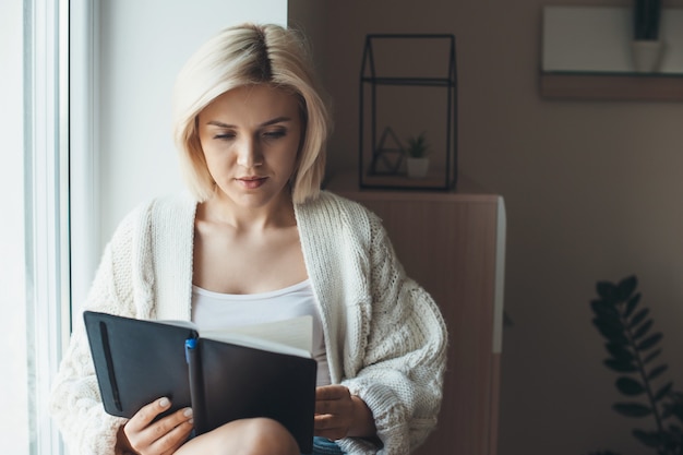 Mujer rubia con un suéter de punto está leyendo un libro sentado cerca de la ventana en casa