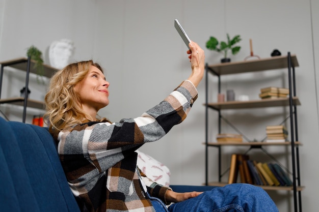 Foto mujer rubia tomando una selfie en el teléfono móvil mientras está sentada en el sofá de la sala de estar