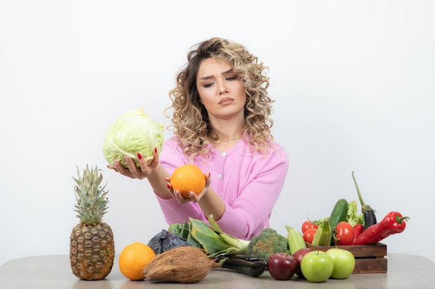 mujer rubia en top rosa con repollo y naranja en la mesa.