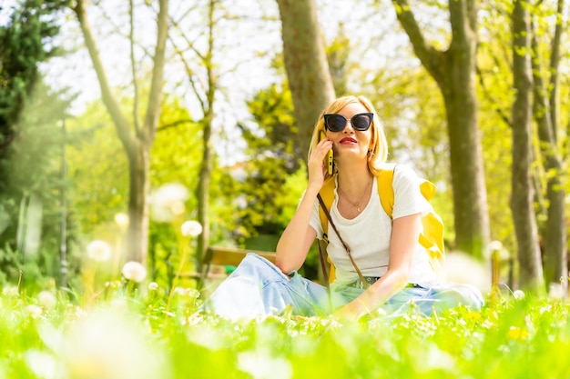 Mujer rubia turista llamando a sus amigas con teléfono con sombrero y gafas de sol sentadas en el césped en primavera junto a margaritas en un parque de la ciudad