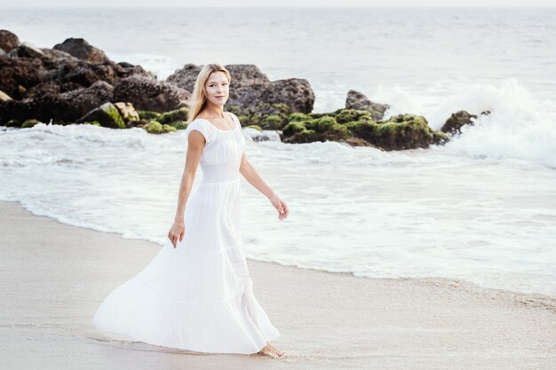 Mujer rubia turística madura feliz atractiva en vestido blanco largo en la playa tropical de la arena asiática.