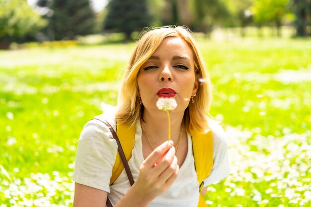 Una mujer rubia turística que sopla una planta de diente de león con un sombrero y gafas de sol sentada en el césped en primavera junto a las margaritas en un parque de la ciudad