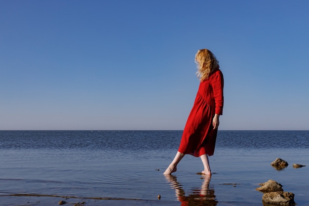 Mujer rubia con un vestido largo de lino rojo de pie en el mar por la mañana.