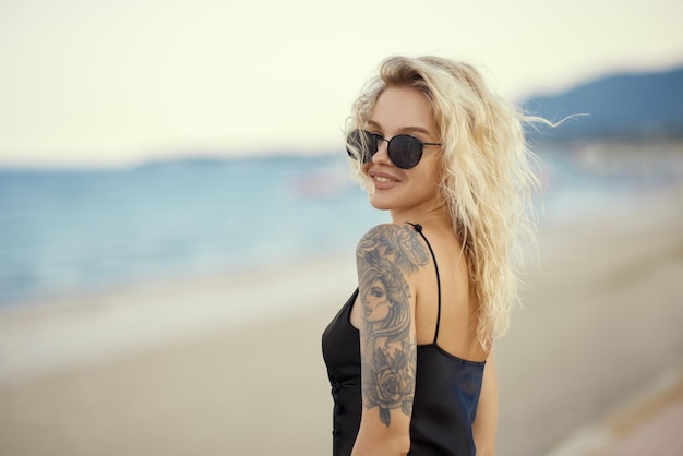 Mujer rubia con vestido negro al aire libre contra la orilla del mar en el fondo durante la playa de verano