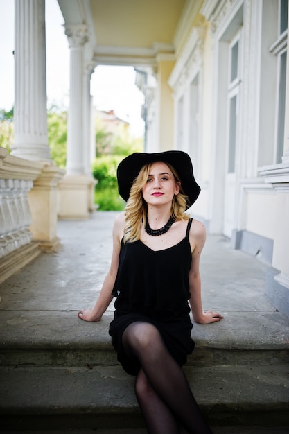 Mujer rubia en vestido negro, collares y sombrero contra casa vintage.