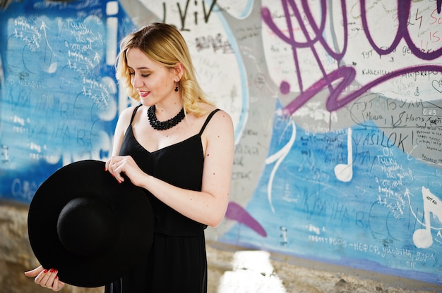 Mujer rubia en vestido negro, collares y sombrero contra la pared de graffiti.