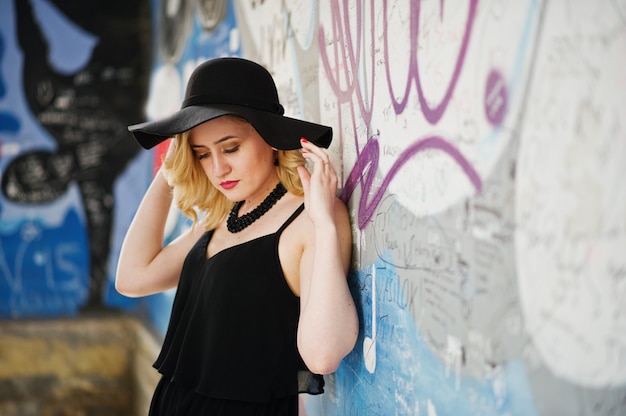 Mujer rubia en vestido negro, collares y sombrero contra la pared de graffiti.