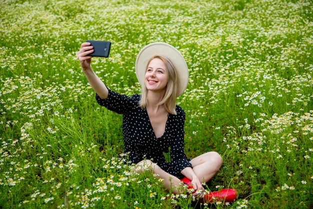 Mujer rubia en vestido negro sentada en campo chamomiles campo de flores y utilizando el teléfono móvil