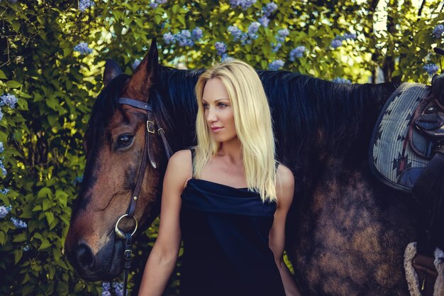 Mujer rubia en vestido de noche negro posando con caballo marrón.