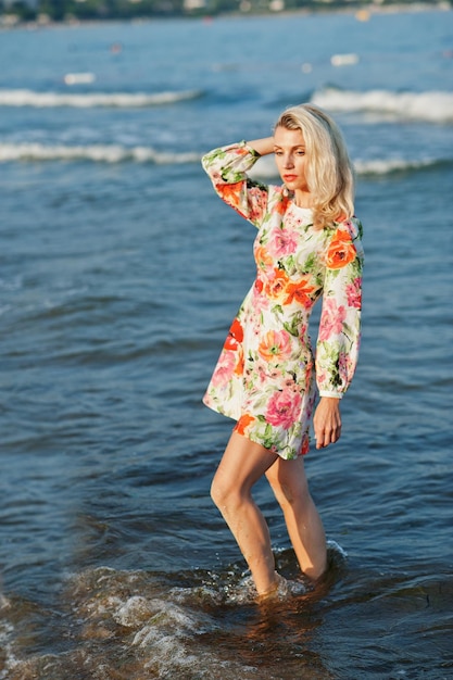 Mujer rubia con vestido de pie en la playa de arena en la orilla del mar disfrutando de la vista del atardecer