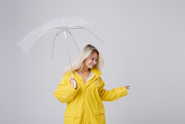 Mujer rubia vistiendo impermeable amarillo con paraguas transparente comprobando el clima si está lloviendo