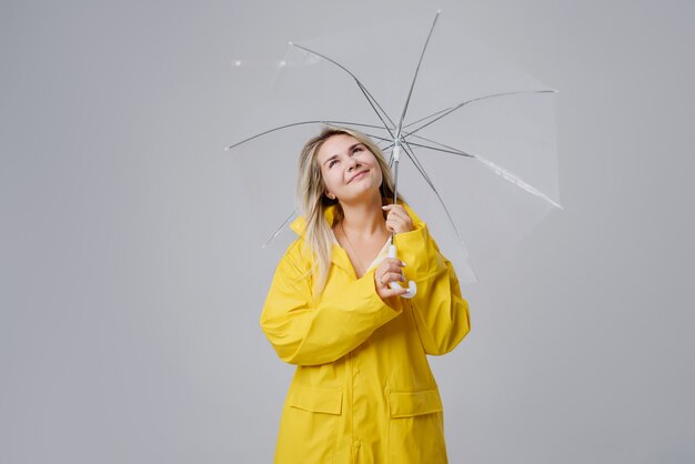 Mujer rubia vistiendo impermeable amarillo con paraguas transparente comprobando el clima si está lloviendo