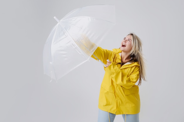 Mujer rubia vistiendo impermeable amarillo con paraguas transparente comprobando el clima si está lloviendo