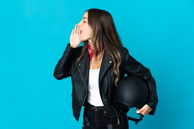 Mujer rumana sosteniendo un casco de motocicleta aislado en la pared azul gritando con la boca abierta hacia el lado