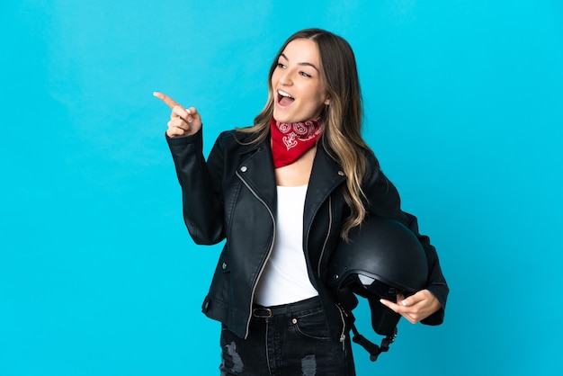 Mujer rumana sosteniendo un casco de motocicleta aislado sobre fondo azul apuntando con el dedo hacia el lado y presentando un producto