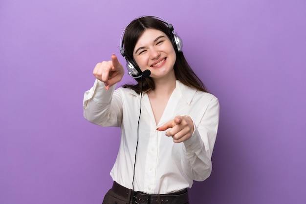 Una mujer rusa televendedora que trabaja con un auricular aislado en un fondo morado te señala con el dedo mientras sonríe