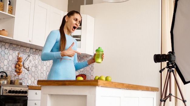 Mujer sana grabando su video blog sobre aditivos alimentarios saludables mientras está de pie en la cocina. Ella sostiene una botella de nutrición deportiva y sonríe.