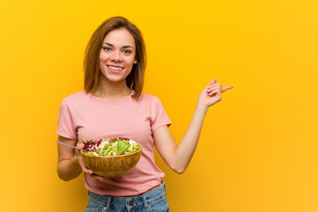 Mujer sana joven que sostiene una ensalada que sonríe alegremente que señala con el índice lejos.