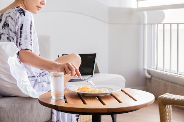 Mujer sana sentada en un sofá comiendo fruta fresca mientras trabaja en una laptop