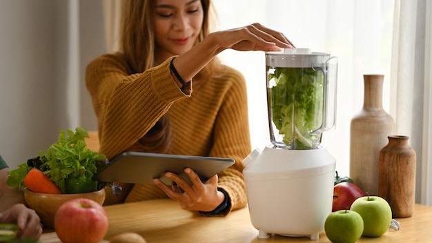 Mujer sana usando tableta digital y haciendo batido de desintoxicación de vegetales verdes en la cocina