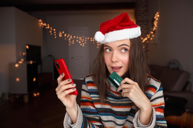 Mujer en santa helper hat con teléfono inteligente y tarjeta de crédito mirando a otro lado