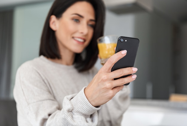 Mujer satisfecha de 30 años bebiendo jugo de naranja y usando un teléfono móvil, mientras descansa en una habitación moderna y luminosa