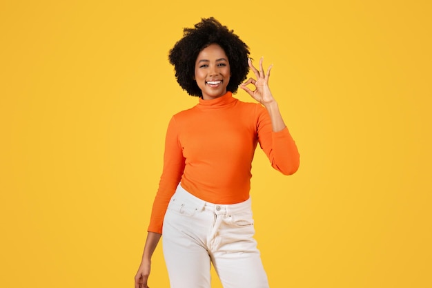 Una mujer satisfecha con cabello afro en cuello atornillado naranja y pantalones blancos hace la señal de OK