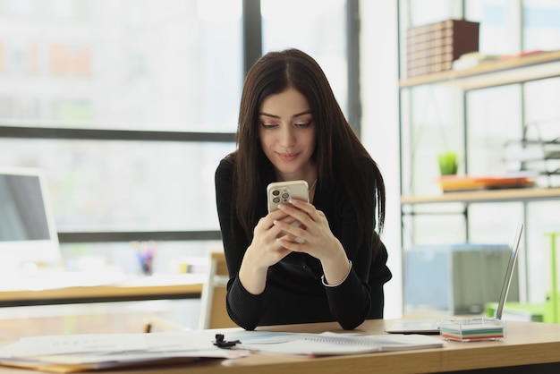 Mujer satisfecha envía mensajes a su novio a través de un teléfono inteligente sentado en la mesa en la oficina morena