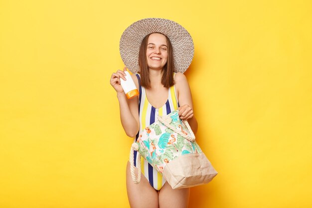 Mujer satisfecha con traje de baño aislado en fondo amarillo de pie con bolsa de playa en sombrero de paja sosteniendo su crema protectora spf disfrutando de la calidad